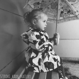 Katarzyna Rudowiecka - Czarno-białe zdjęcie dziewczynki z kucykiem, siedzącej na schodach z parasolem w dłoni, ubrana w kurtkę z motywem zwierzęcym.