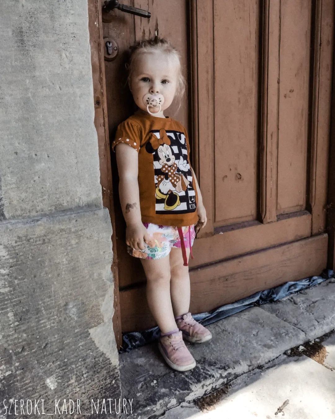 Dziewczynka z blond włosami i smoczkiem w ustach, oparta o drewniane drzwi i kamienną ścianę; ubrana w pomarańczową koszulkę z Minie i wzorzyste spodenki.
