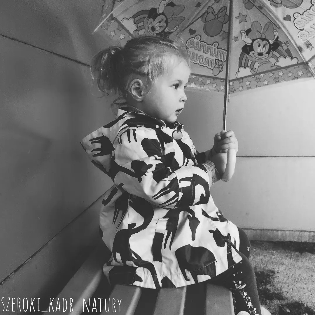 Czarno-białe zdjęcie dziewczynki z kucykiem, siedzącej na schodach z parasolem w dłoni, ubrana w kurtkę z motywem zwierzęcym.