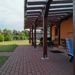 Zadaszona altana z ciemnego drewna, z przezroczystym dachem, przylegająca do pomarańczowego budynku. Widoczny brukowany chodnik, trawnik i ogrodzenie w tle.