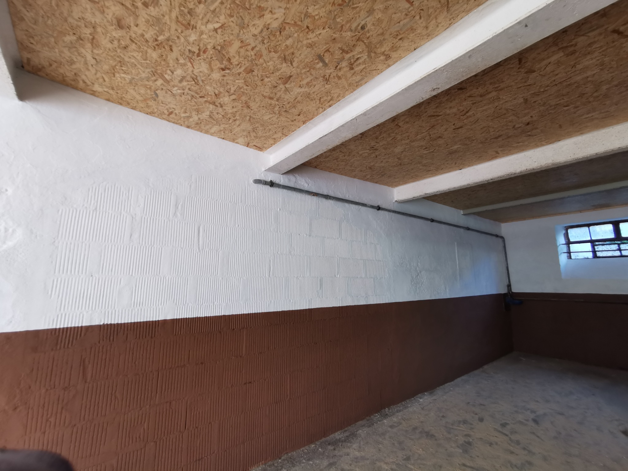 Surowe wnętrze z wykończonym stropem z płyty OSB, dwukolorowe ściany z widoczną fakturą i rurą instalacyjną pod sufitem, okno w tle.
