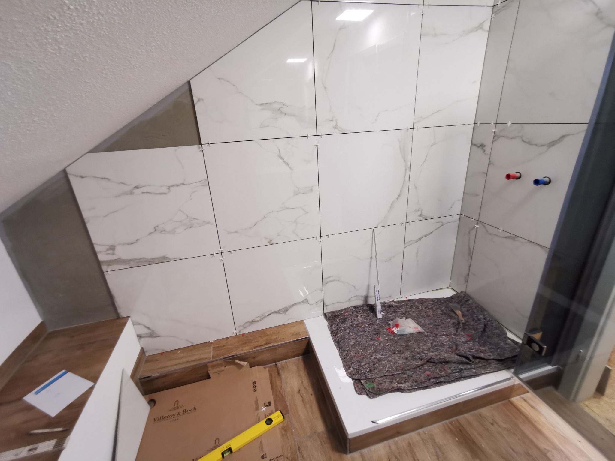 Montaż płytek imitujących marmur w łazience na poddaszu, widoczne krzyżyki dystansowe, poziomica i karton Villeroy & Boch.