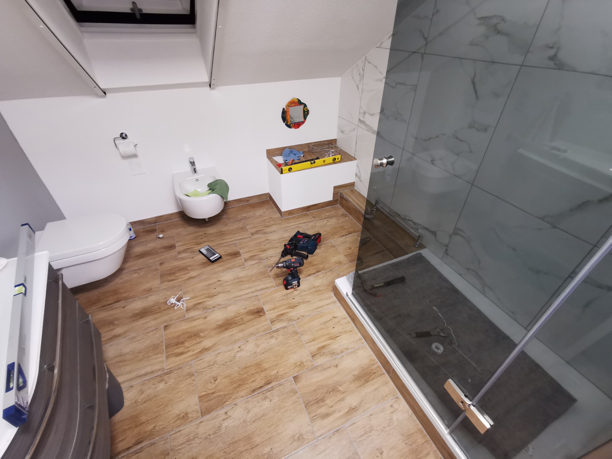 Montaż kabiny prysznicowej w łazience na poddaszu: widoczne narzędzia, poziomica, płytki imitujące marmur, umywalka ze wzorem ryby na ścianie.