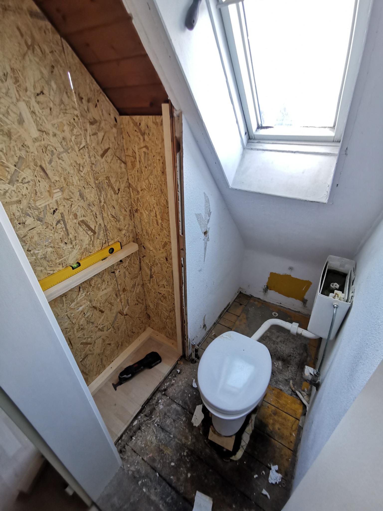 Wnętrze małej łazienki na poddaszu w trakcie remontu; widoczna toaleta, okno dachowe, ściany przygotowane do wykończenia oraz fragment konstrukcji z płyty OSB z poziomicą i narzędziem wielofunkcyjnym.