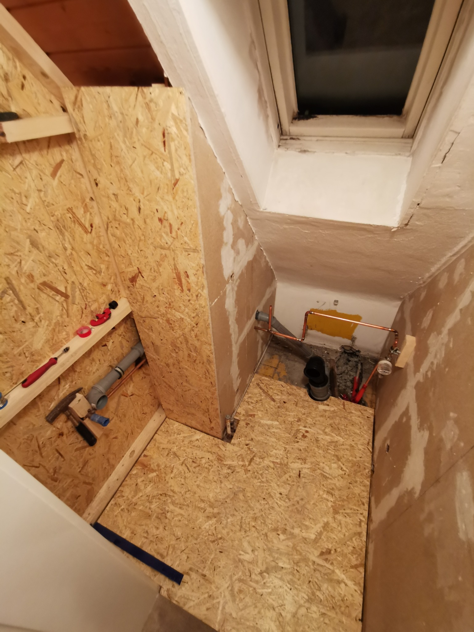 Wnętrze w trakcie budowy, z widoczną instalacją hydrauliczną, drewnianą konstrukcją obudowy i oknem dachowym.