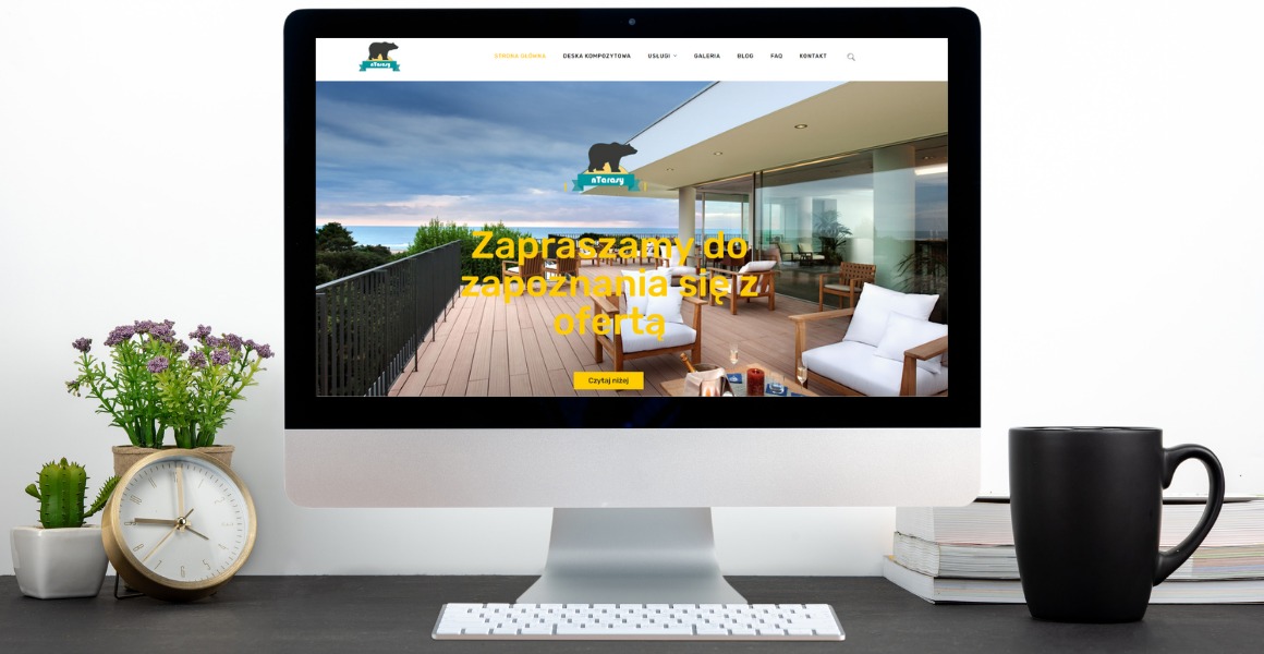 Ekran komputera iMac wyświetlający stronę internetową ntarasy.pl z widokiem na taras z desek kompozytowych, w tle morze i roślinność; na biurku obok monitora zegar, roślina w doniczce i czarny kubek.