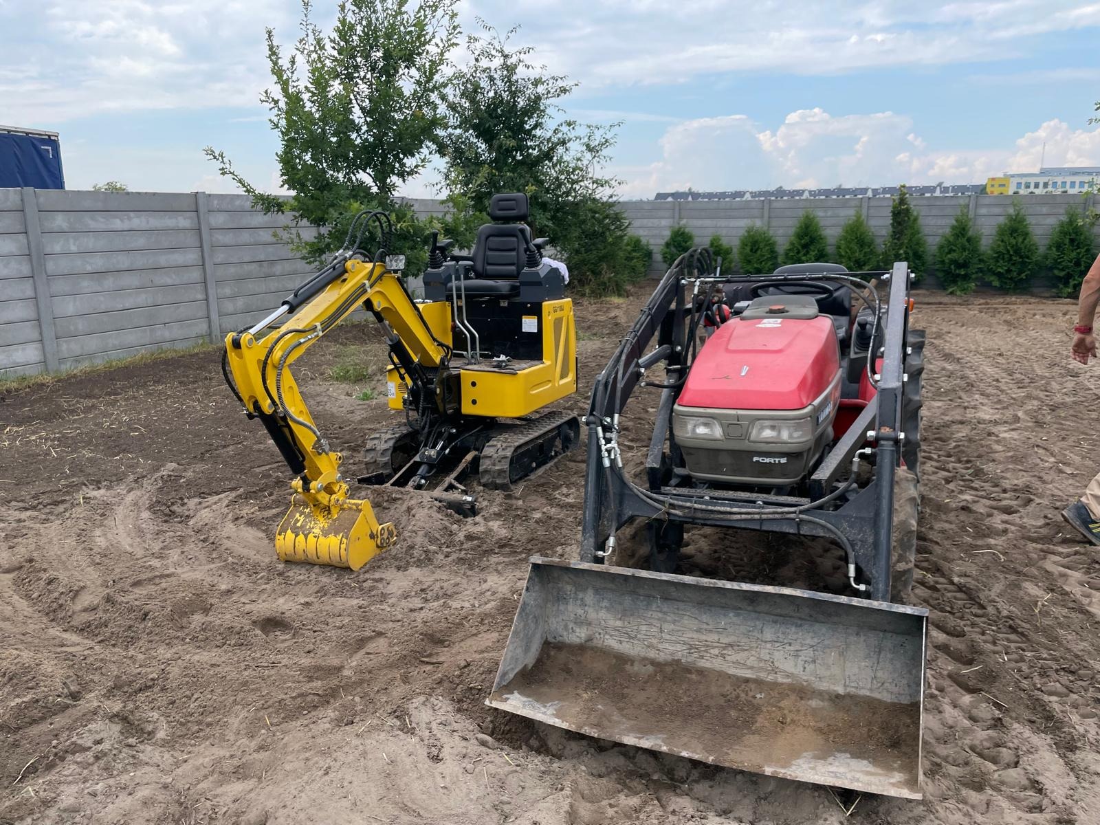 Żółta mini koparka i czerwony traktor z łyżką na nierównym, przygotowanym terenie, w tle betonowy płot i drzewa.