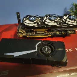 Dwie karty graficzne: MSI z turbinowym chłodzeniem i Arctic z trzema wentylatorami, leżące na pudełku po płycie głównej ASUS.