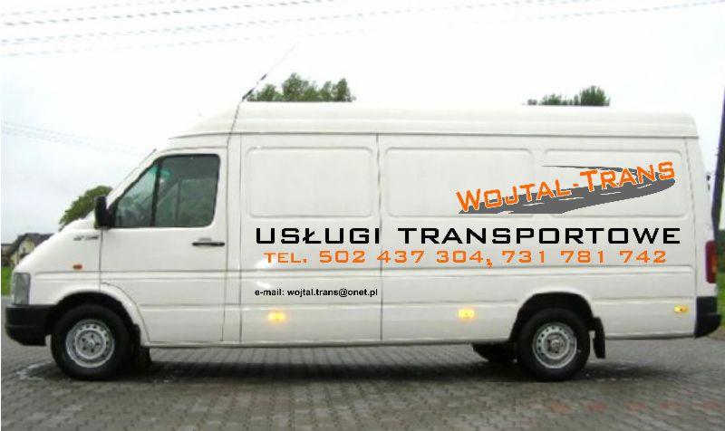 Biały bus z napisem 'WOJTAL-TRANS' i danymi kontaktowymi, zaparkowany na zewnątrz. Widok z boku, na tle zabudowań.