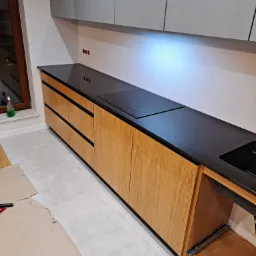 Nowoczesna kuchnia z drewnianymi szafkami i czarnym blatem, widoczna płyta indukcyjna i zlewozmywak, częściowo zakryta podłoga podczas montażu.