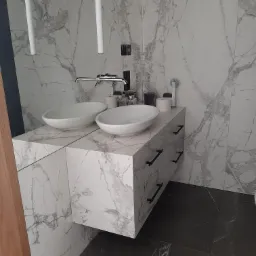 Nowoczesna łazienka z dwoma owalnymi umywalkami nablatowymi na szafce z szufladami, wykończona płytami imitującymi marmur, z minimalistyczną armaturą i oświetleniem.