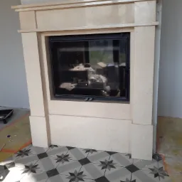 Beżowy obudowany wkład kominkowy z kamienia, czarny front, płytki podłogowe w gwiazdy, widoczne ślady prac wykończeniowych.