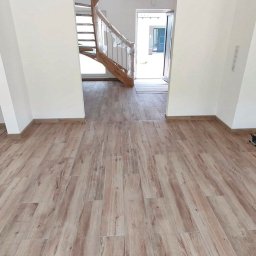 Dobrafirma PL Usługi budowlane Wadim Bagiński - Jasna podłoga z paneli imitujących drewno, świeżo położona w nowym domu, widok na schody z drewnianą barierką zabezpieczoną folią i otwarte drzwi wejściowe.