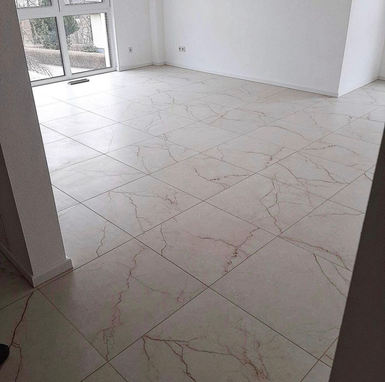 Jasne, marmurowe płytki podłogowe ułożone w kwadraty w nowo wykończonym pokoju z dużym oknem i białymi ścianami.