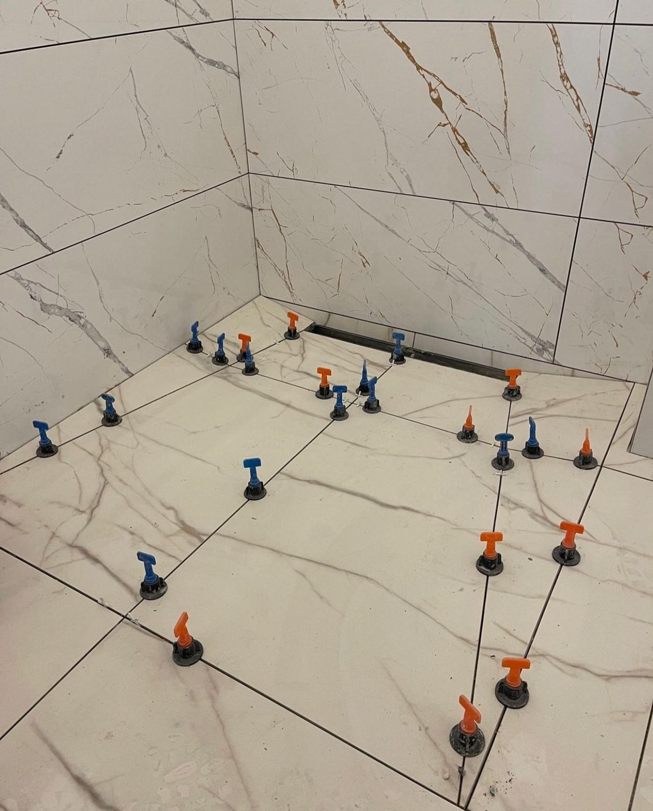 Podłoga w trakcie układania płytek imitujących marmur, z widocznymi systemami poziomowania w kolorach niebieskim i pomarańczowym, oraz odpływem liniowym w rogu pomieszczenia.