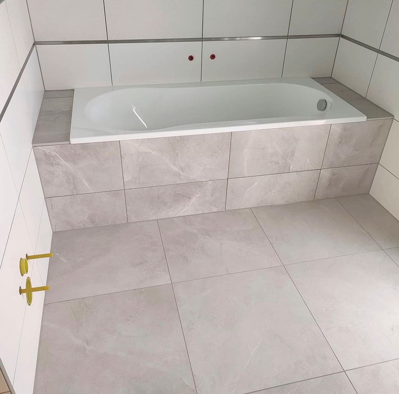 Wanna obudowana płytkami imitującymi marmur, z widocznymi złotymi przyłączami do baterii, na tle białych płytek ściennych z metalową listwą dekoracyjną.