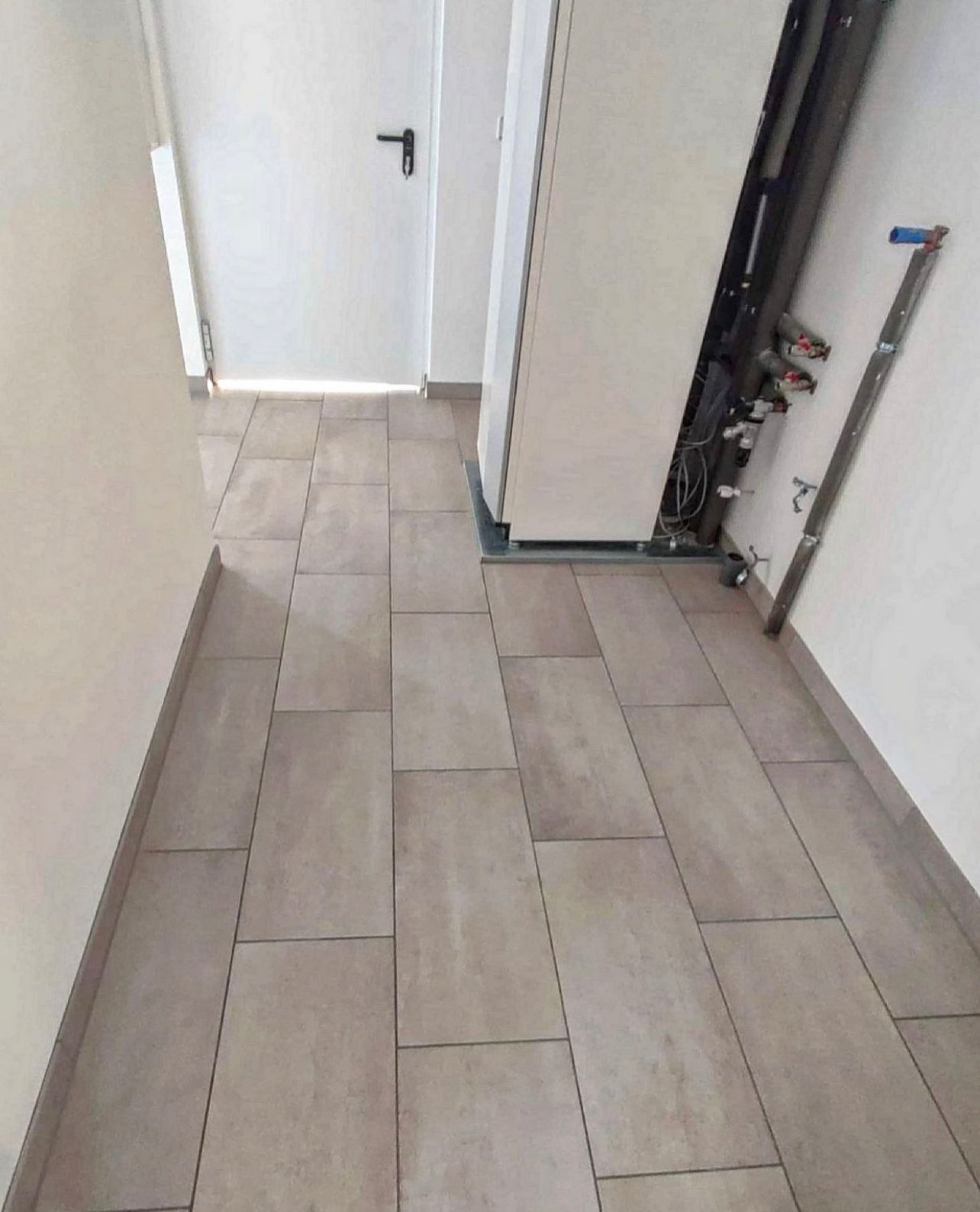 Beżowe, prostokątne płytki podłogowe ułożone w korytarzu, widoczne rury instalacji grzewczej przy ścianie