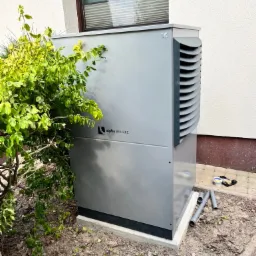 Szara pompa ciepła Alpha Innotec na betonowej podstawie, obok budynku z oknem, z widocznymi rurami i narzędziami na ziemi.