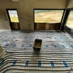 New Era Energy - Montaż rur grzewczych na folii z nadrukiem w surowym wnętrzu z widokiem na budowę przez okna.