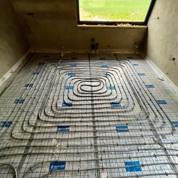 New Era Energy - Rozłożone rury systemu grzewczego podłogowego na izolacji z siatką, widoczne okno i surowe ściany w trakcie prac wykończeniowych.