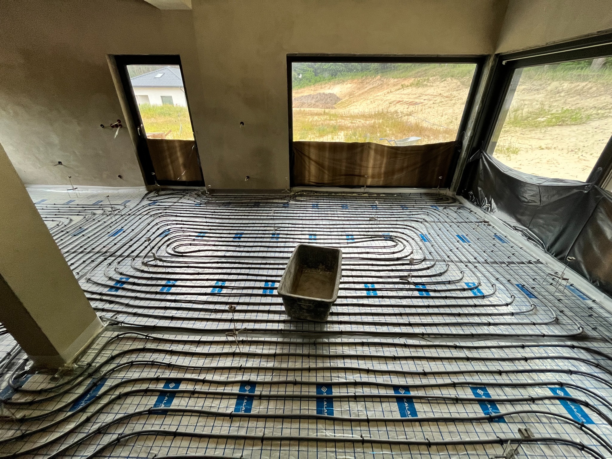 Montaż rur grzewczych na folii z nadrukiem w surowym wnętrzu z widokiem na budowę przez okna.