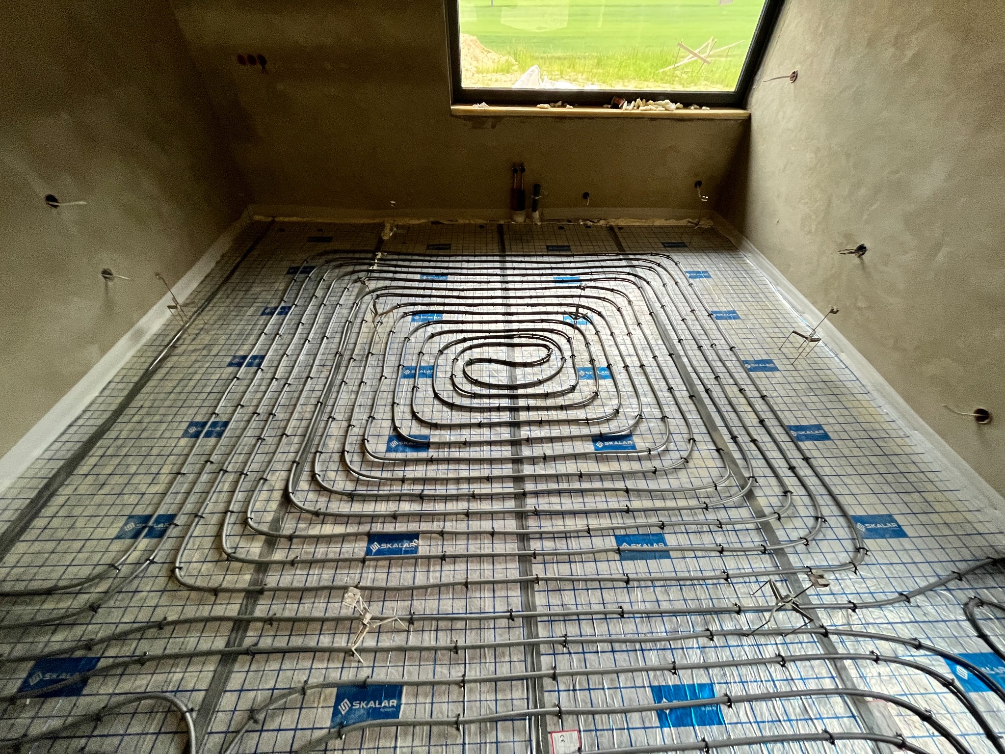 Rozłożone rury systemu grzewczego podłogowego na izolacji z siatką, widoczne okno i surowe ściany w trakcie prac wykończeniowych.