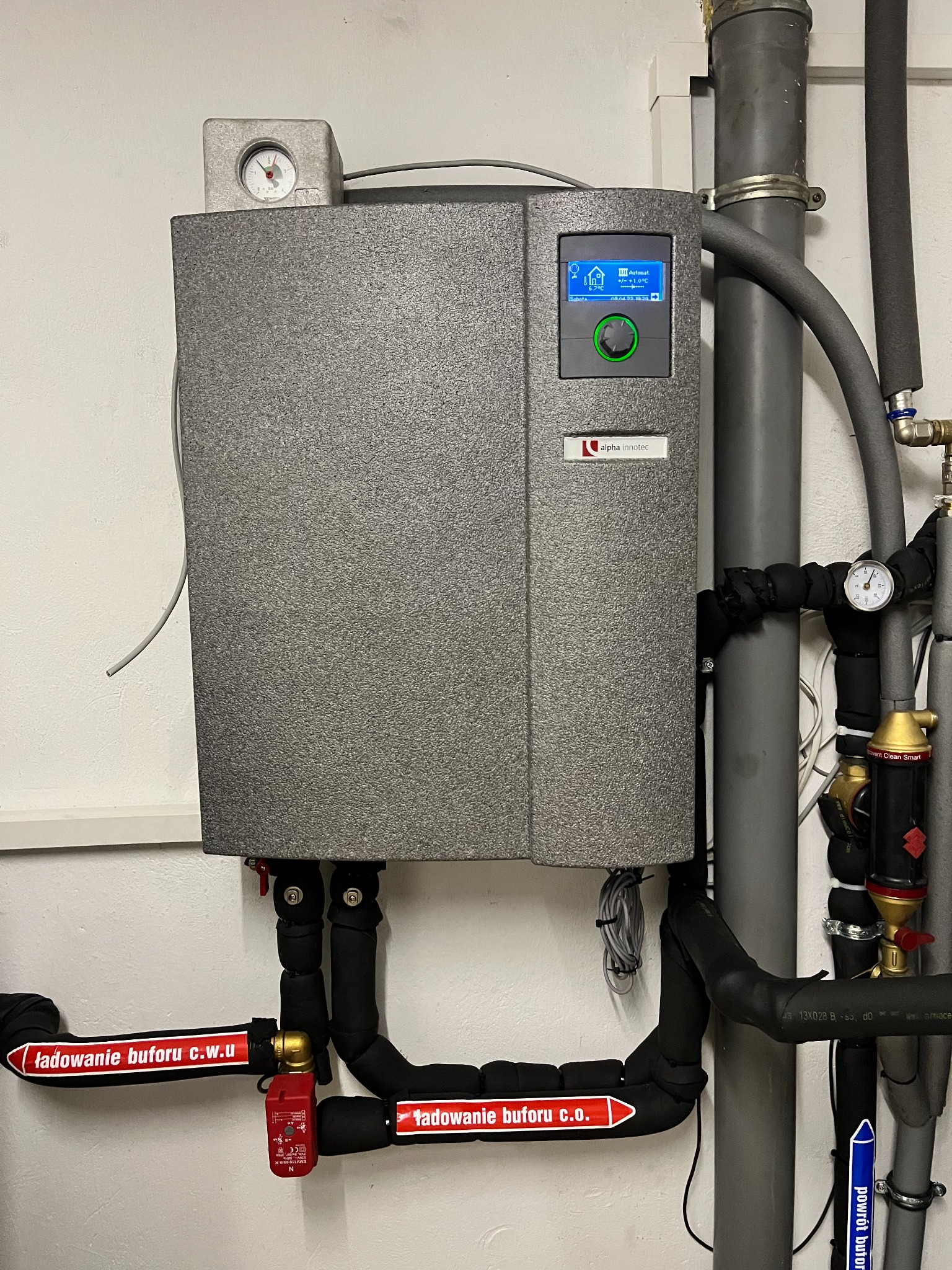 Szara jednostka centralna z wyświetlaczem i logiem 'alpha innotec', podłączona do rur z izolacją i zaworów, z etykietami 'ładowanie buforu c.w.u.' i 'ładowanie buforu c.o.' oraz 'powrót buforu'.