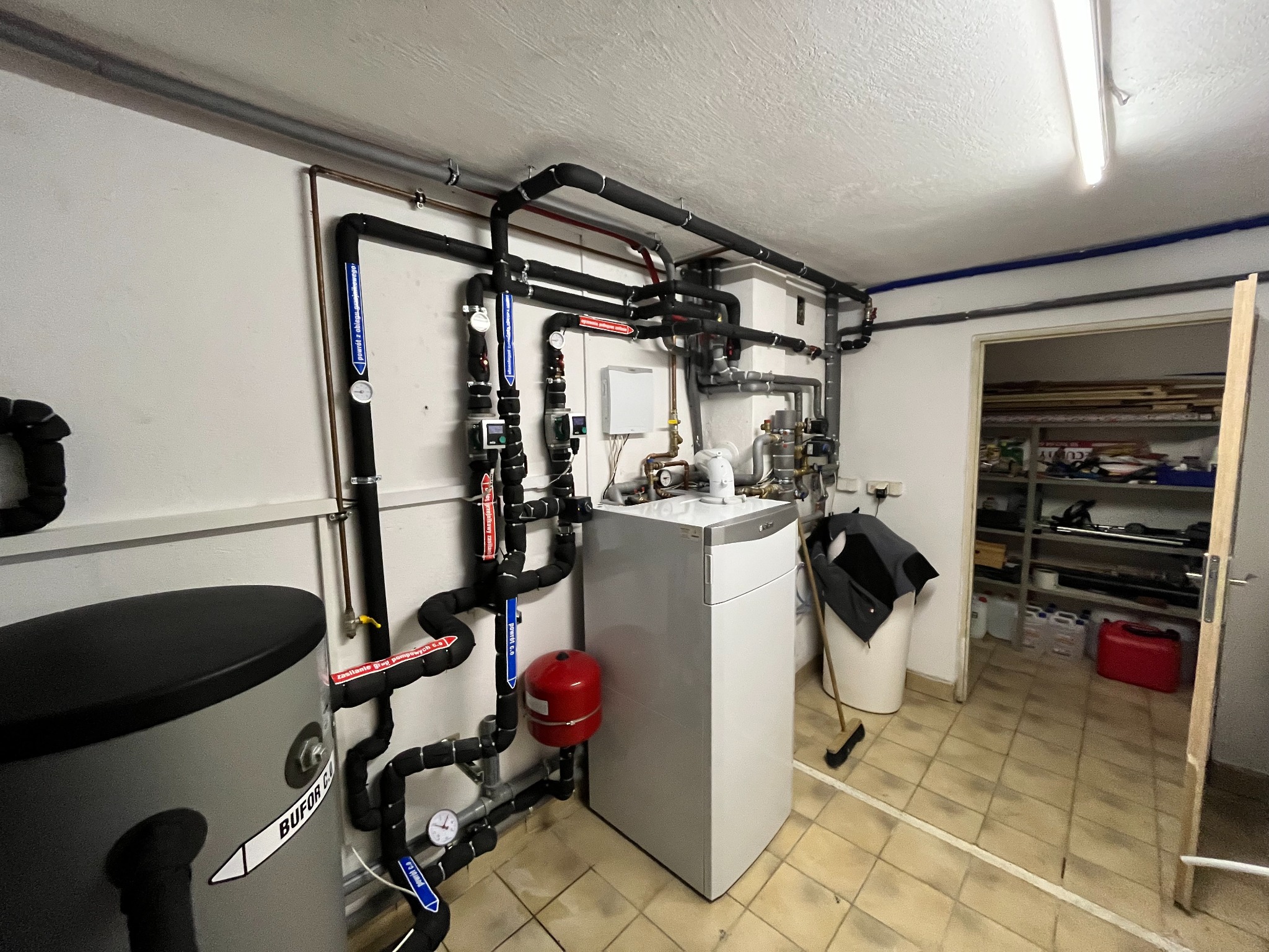 Zainstalowana kotłownia z szarym zbiornikiem buforowym marki BUFOR C.O., białym kotłem i licznymi rurami z izolacją, manometrami i zaworami w pomieszczeniu technicznym z płytkami na podłodze...