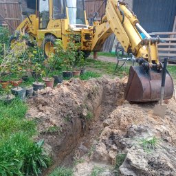 JC Czosnkowe Gospodarstwo Rolne Caroline Dziwir - Żółta koparko-ładowarka Komatsu przy wykopanym wąskim rowie w ziemi, obok roślin w donicach i łopaty wbitej w pryzmę ziemi.