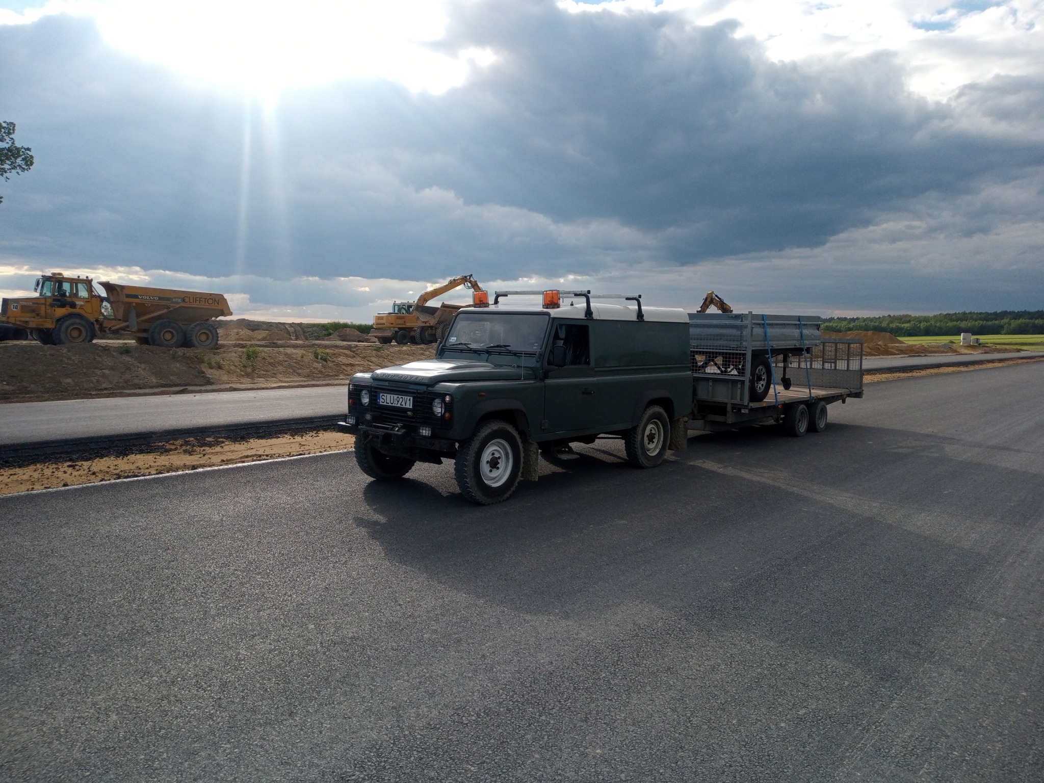 Ciemnozielony Land Rover Defender z przyczepą na nowo wyasfaltowanej drodze; w tle ciężarówki i koparki na placu budowy pod pochmurnym niebem.