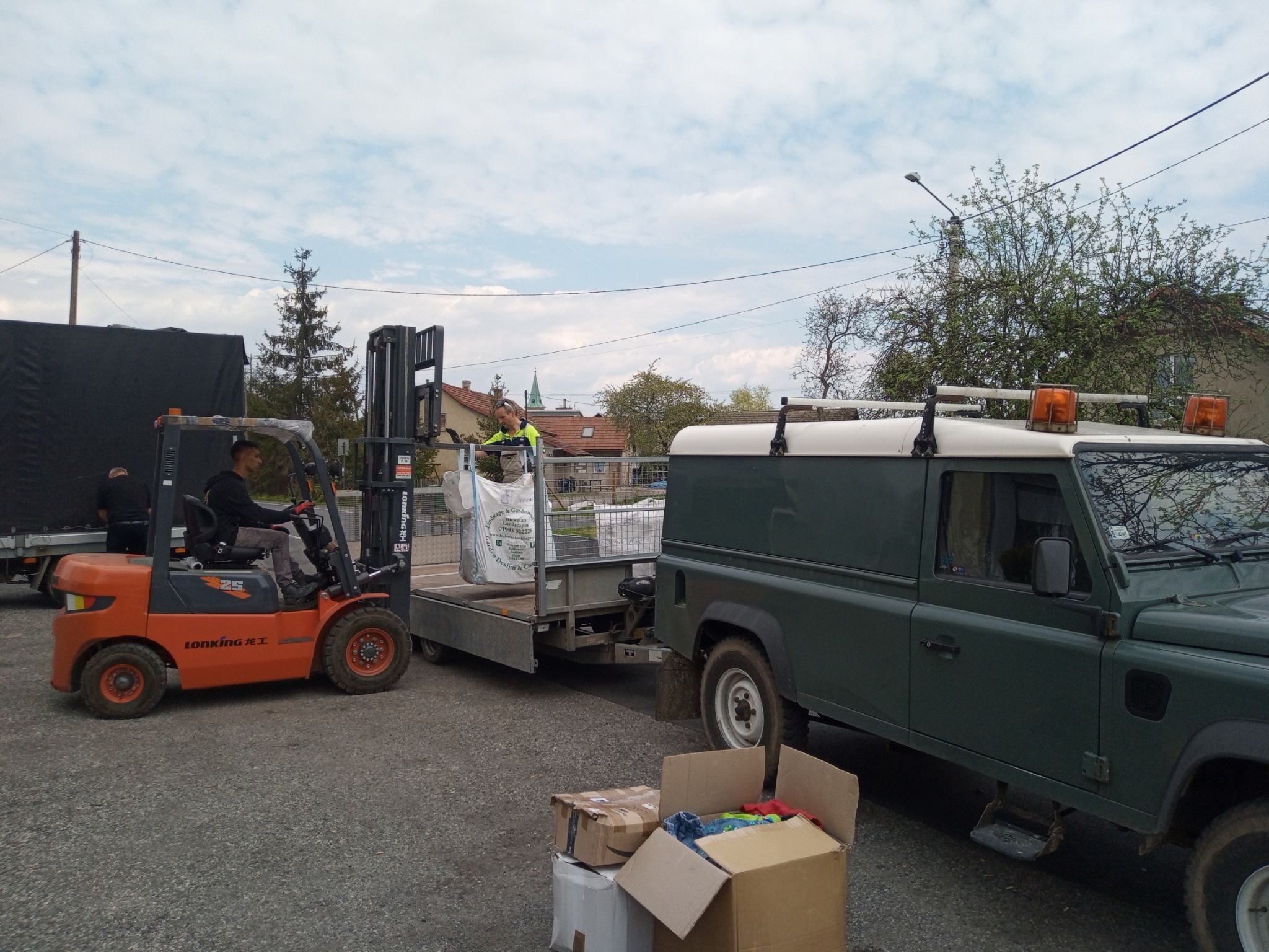 Pomarańczowy wózek widłowy Lonking podnosi biały worek z materiałem na przyczepę ciągniętą przez zielony samochód terenowy, w tle budynki mieszkalne i pochmurne niebo.