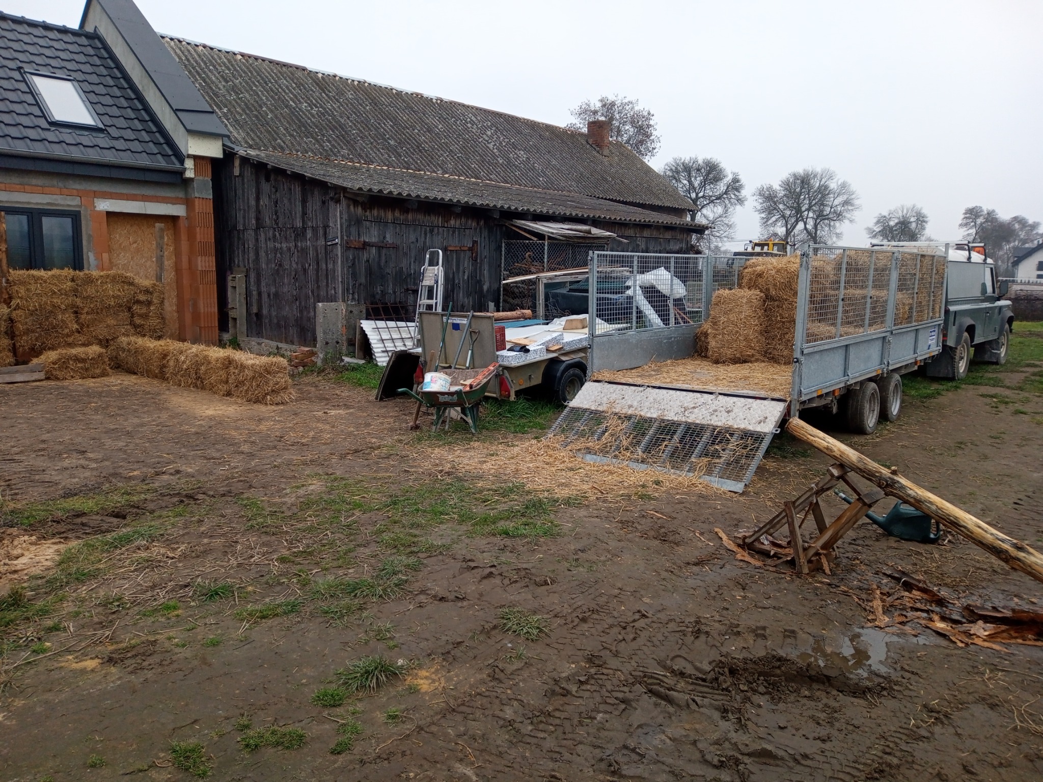 Przygotowany do transportu ładunek sprasowanej słomy na przyczepie za samochodem terenowym, na podwórku obok starego drewnianego budynku gospodarczego i nowego domu w trakcie budowy.