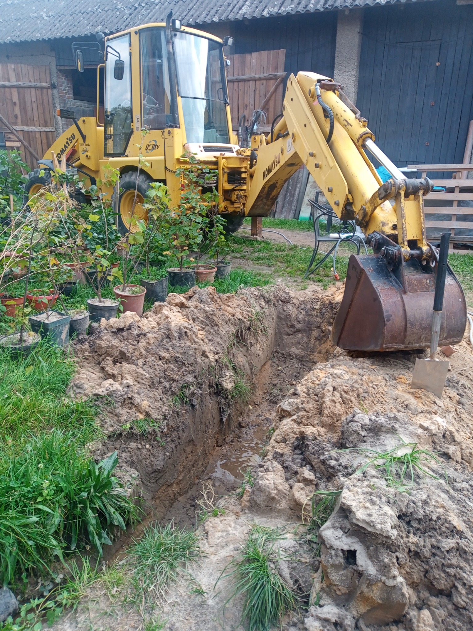 Żółta koparko-ładowarka Komatsu przy wykopanym wąskim rowie w ziemi, obok roślin w donicach i łopaty wbitej w pryzmę ziemi.