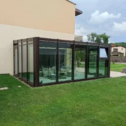 Szklana oranżeria z ciemną ramą przylegająca do domu, widok z zewnątrz na trawnik, wnętrze w trakcie wykańczania.