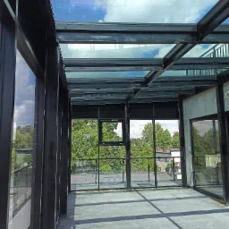 Szklany dach i ściany oranżerii z czarną stalową konstrukcją, widok na zieleń drzew i błękitne niebo z chmurami.