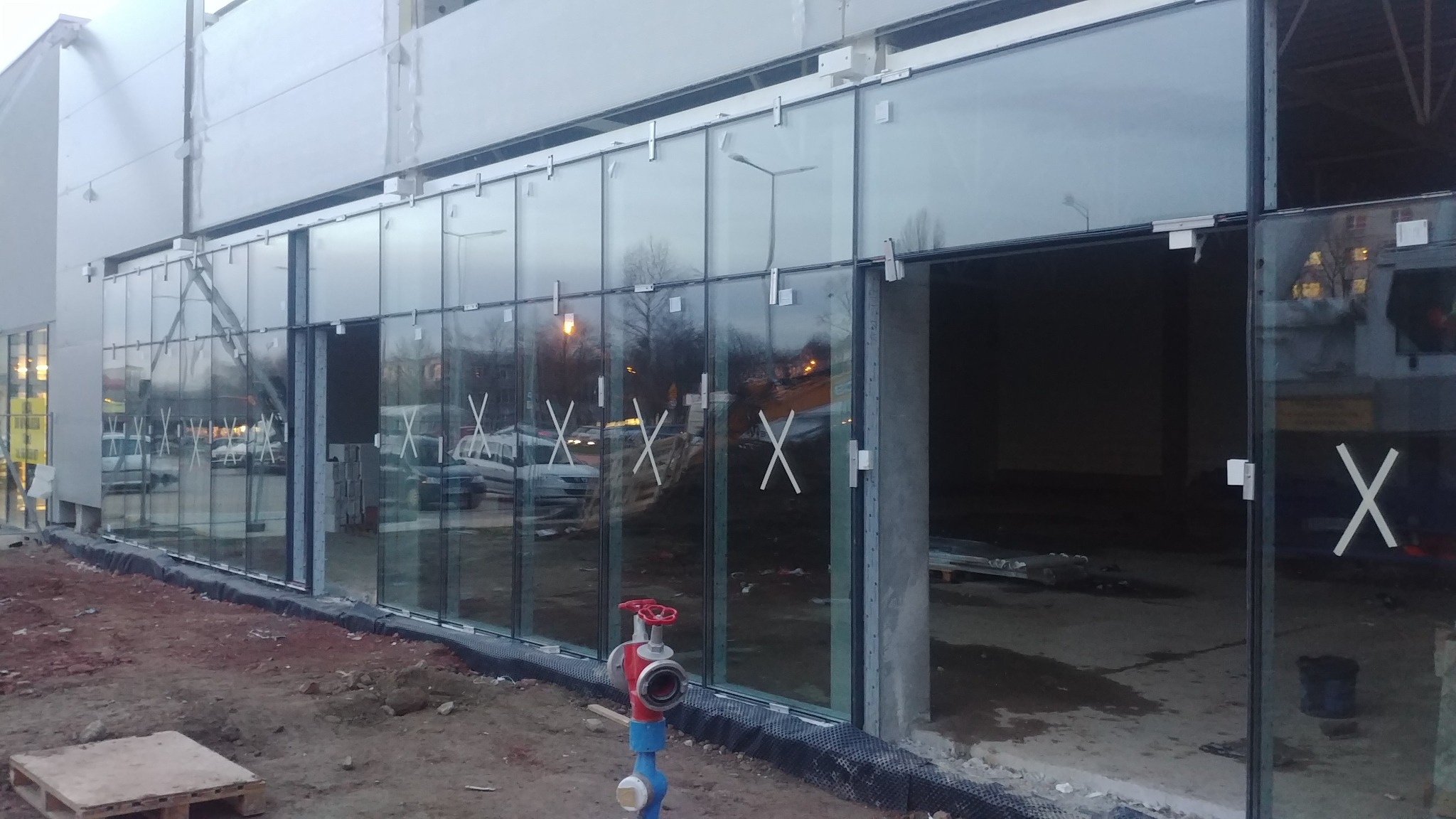 Szklana fasada budynku w trakcie montażu, widoczne mocowania, zabezpieczenia w kształcie X, hydrant i teren budowy.