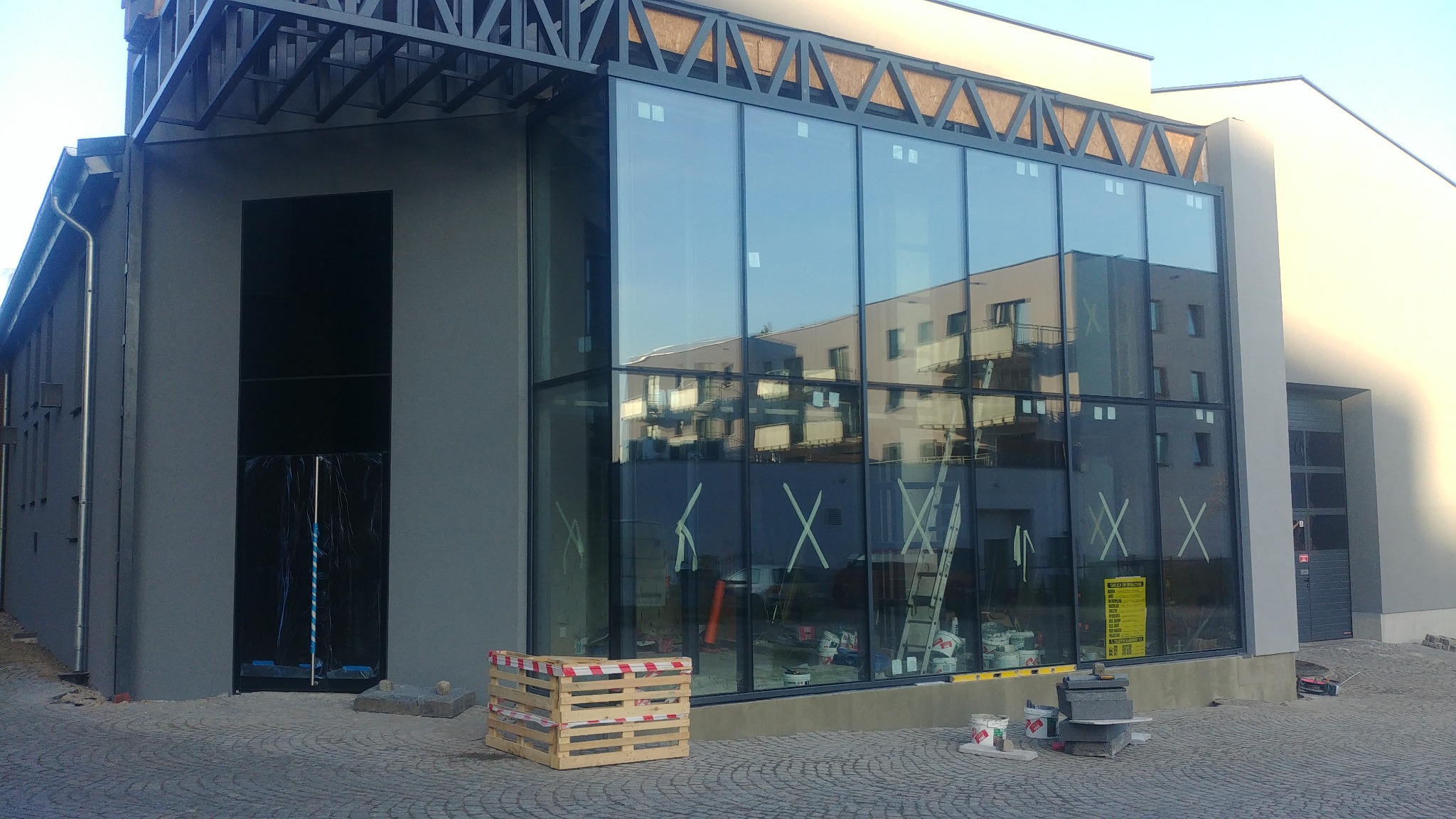 Nowoczesny budynek z dużą szklaną fasadą w trakcie budowy, widoczne rusztowania wewnątrz, paleta i materiały budowlane na zewnątrz.