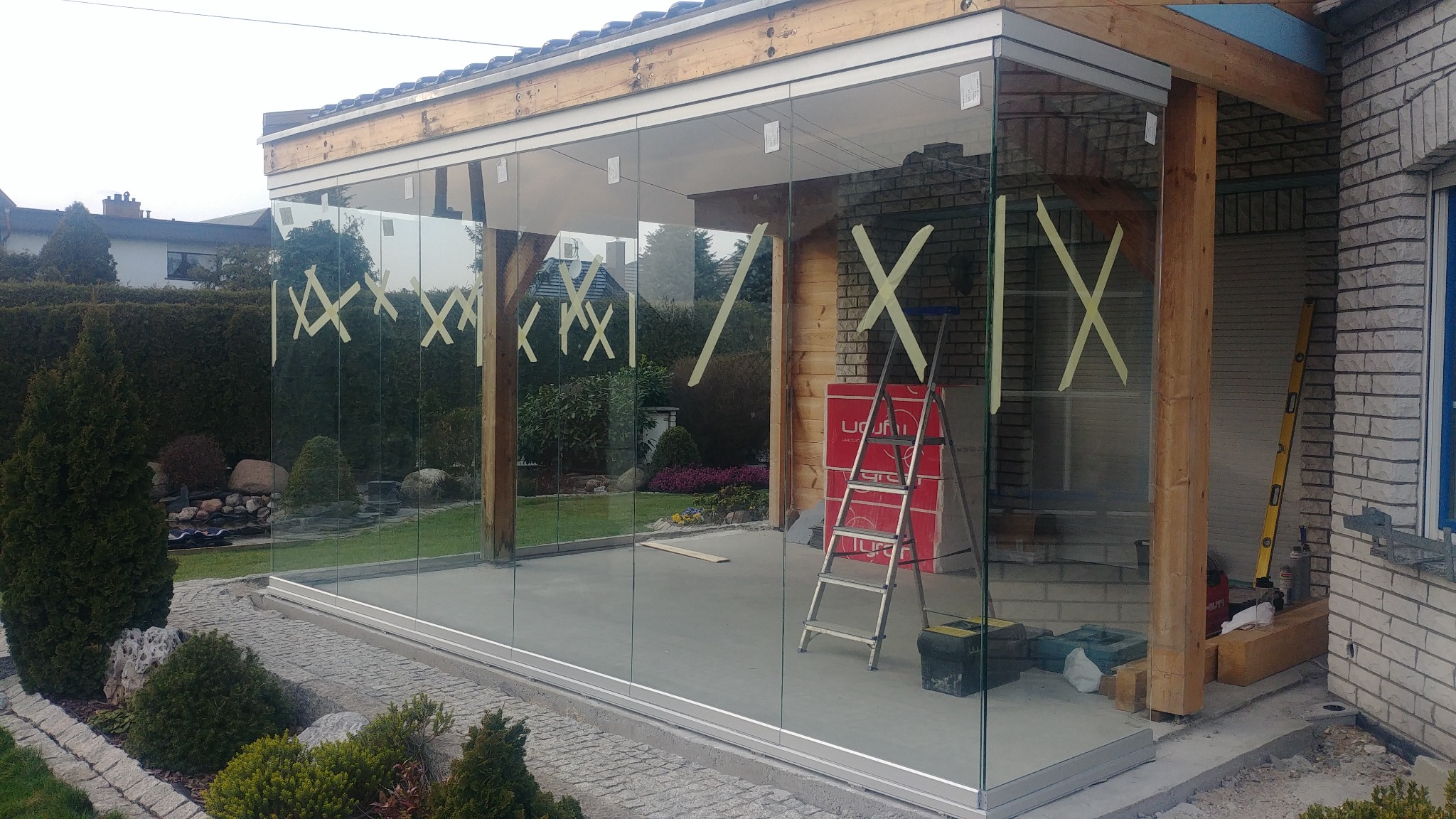 Szklana zabudowa tarasu w trakcie montażu z drewnianą konstrukcją, zabezpieczona taśmą, drabina i narzędzia wewnątrz.