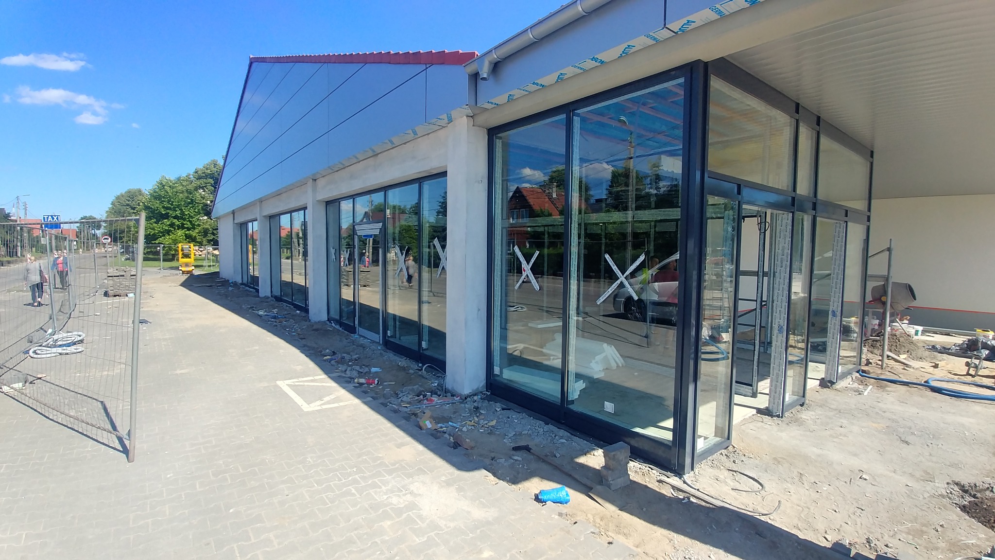 Nowoczesny budynek z dużymi, przeszklonymi oknami w czarnych ramach, widoczne prace wykończeniowe na zewnątrz, elementy rusztowań i piasek.