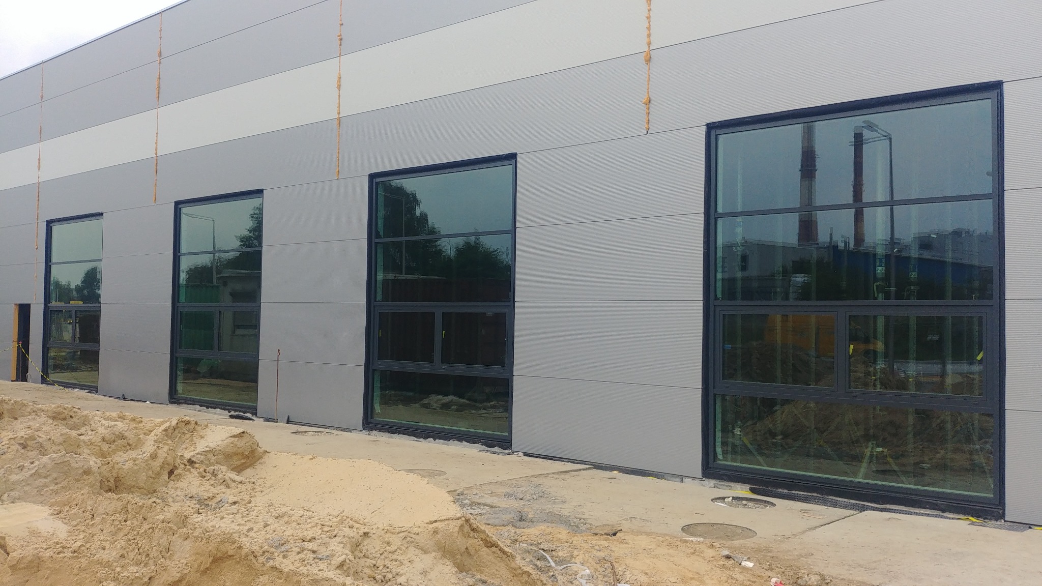 Szara hala przemysłowa z zamontowanymi, dużymi oknami z czarnymi ramami, widoczne prace ziemne na zewnątrz.