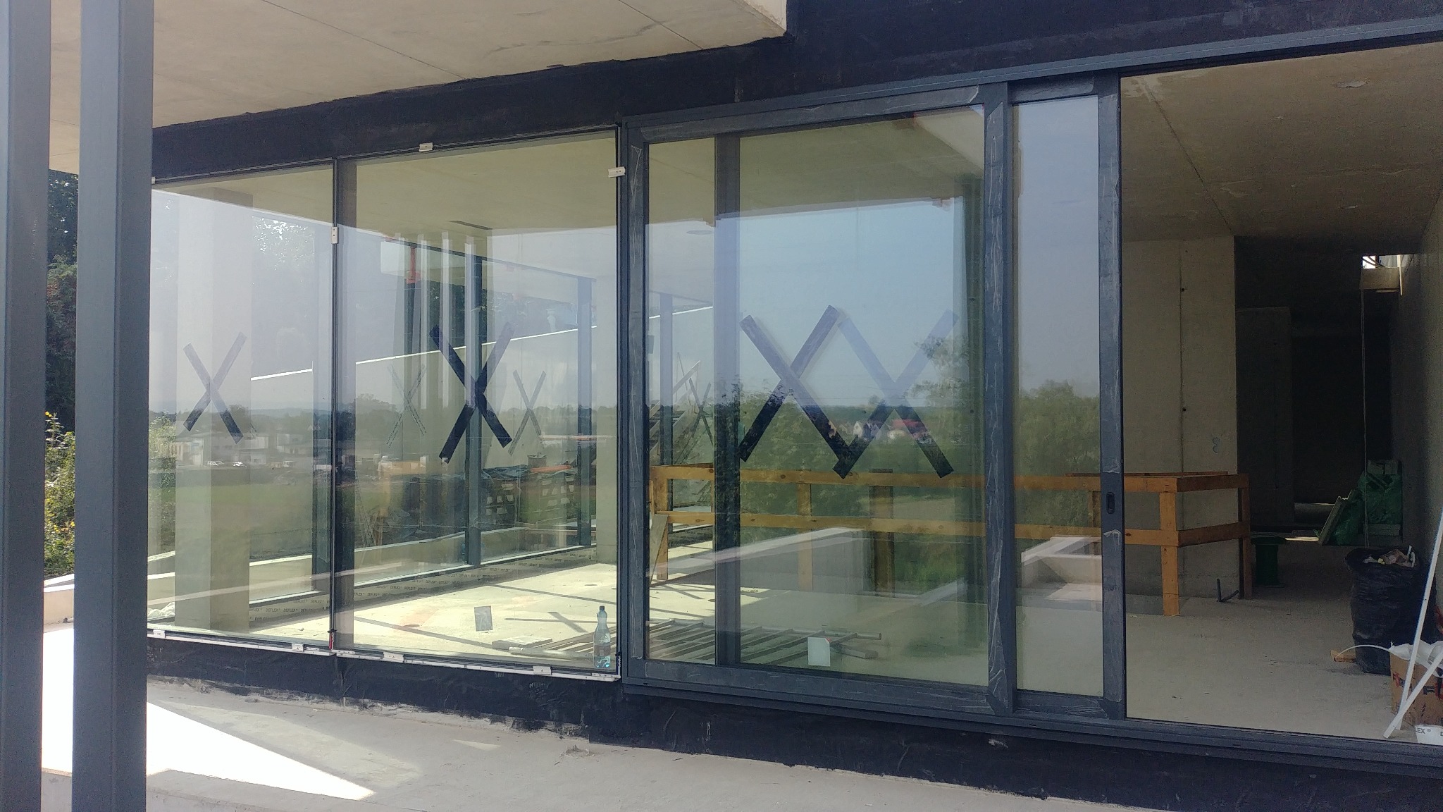 Duże okna przesuwne z czarnymi ramami, zabezpieczone taśmą w kształcie litery X, zamontowane w surowym stanie budynku.