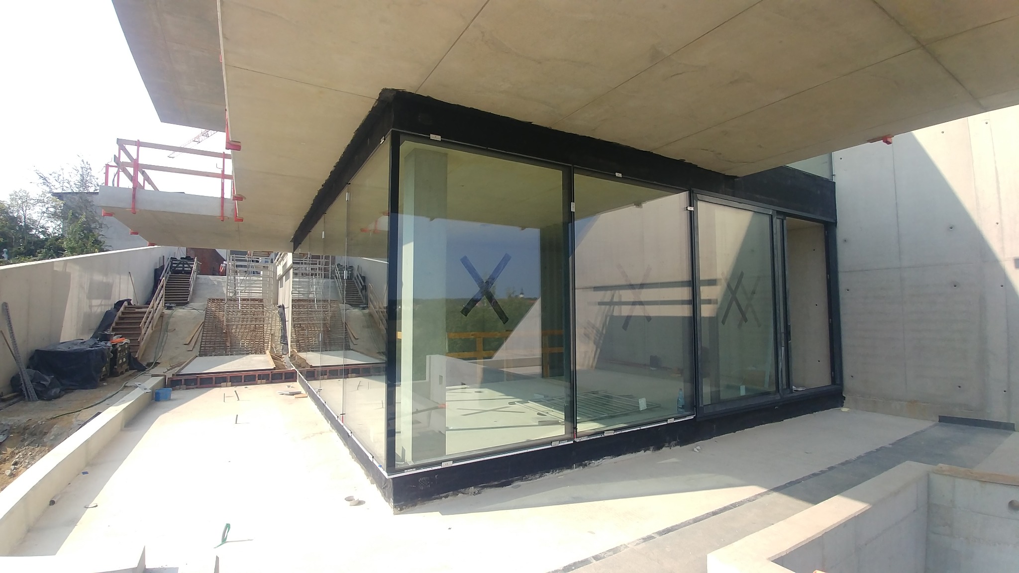 Szklana fasada budynku w trakcie budowy, widoczne betonowe elementy konstrukcyjne i odbicie krajobrazu w szybach.