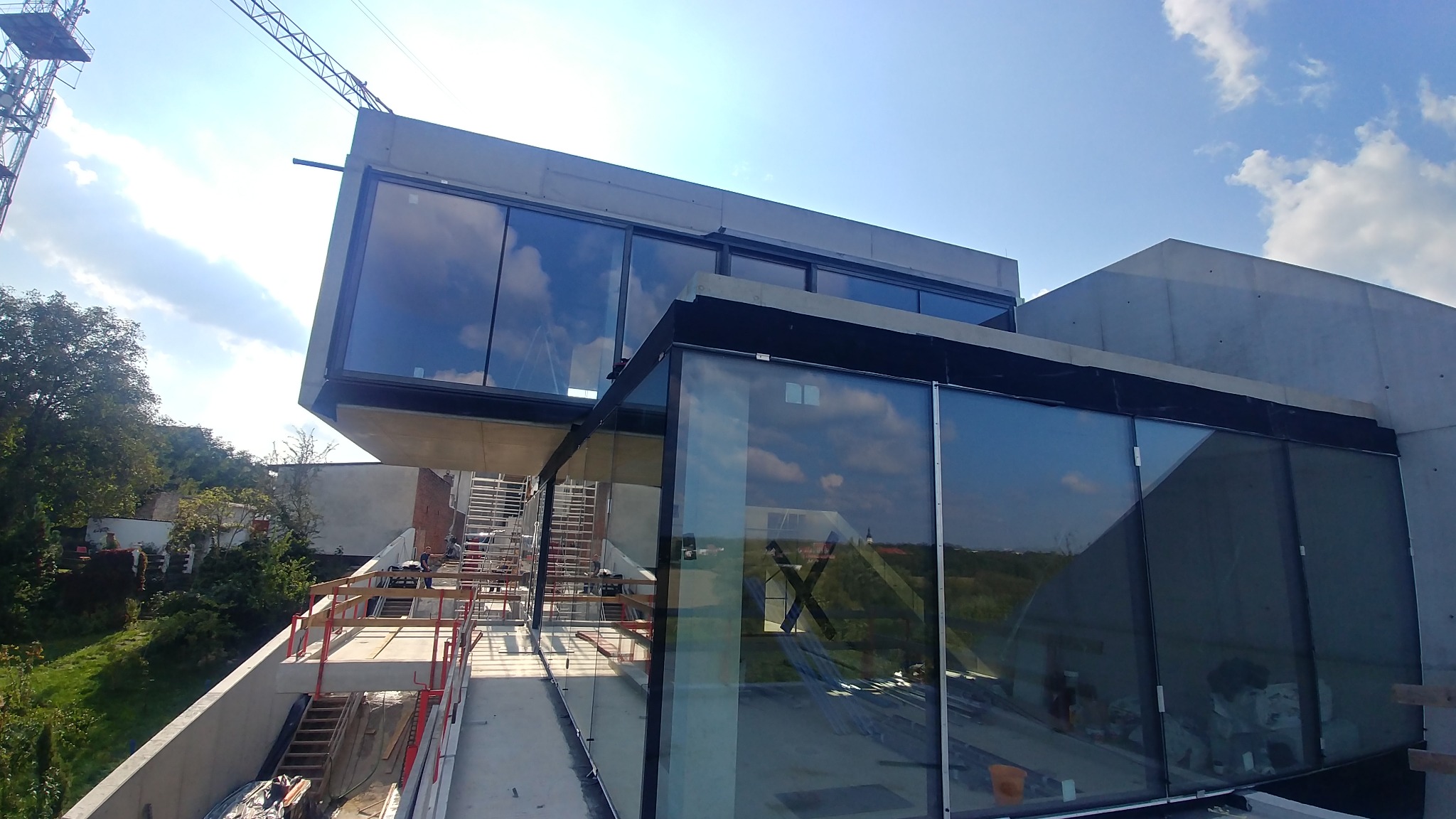 Nowoczesny budynek w trakcie budowy z widocznymi szklanymi fasadami i betonową konstrukcją, z rusztowaniami i żurawiem w tle, na tle błękitnego nieba z chmurami.