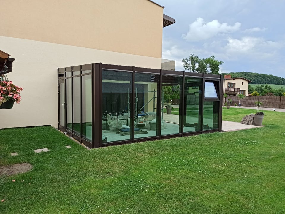 Szklana oranżeria z ciemną ramą przylegająca do domu, widok z zewnątrz na trawnik, wnętrze w trakcie wykańczania.