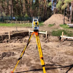 Żółty teodolit ustawiony na trójnogu na placu budowy, w tle wykop i pryzma ziemi.