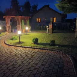 Oświetlona posesja nocą z brukowanym podjazdem, drewnianą altaną, grillem i trawnikiem.