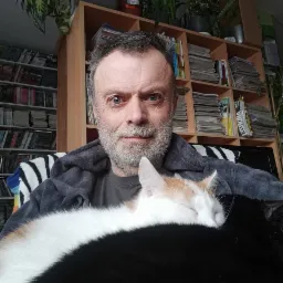 Mężczyzna z siwą brodą i dwoma kotami, jeden śpi na jego ramieniu, a drugi czarny w tle, za nim regał z książkami i roślinami.