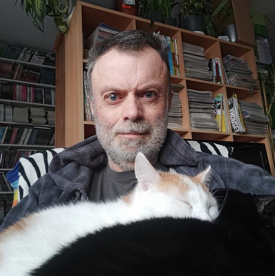 Mężczyzna z siwą brodą i dwoma kotami, jeden śpi na jego ramieniu, a drugi czarny w tle, za nim regał z książkami i roślinami.