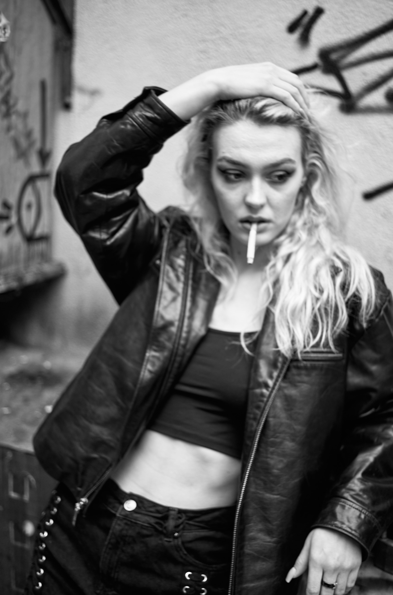 Czarno-białe zdjęcie kobiety w skórzanej kurtce, z papierosem w ustach, stojącej na tle ściany z graffiti.