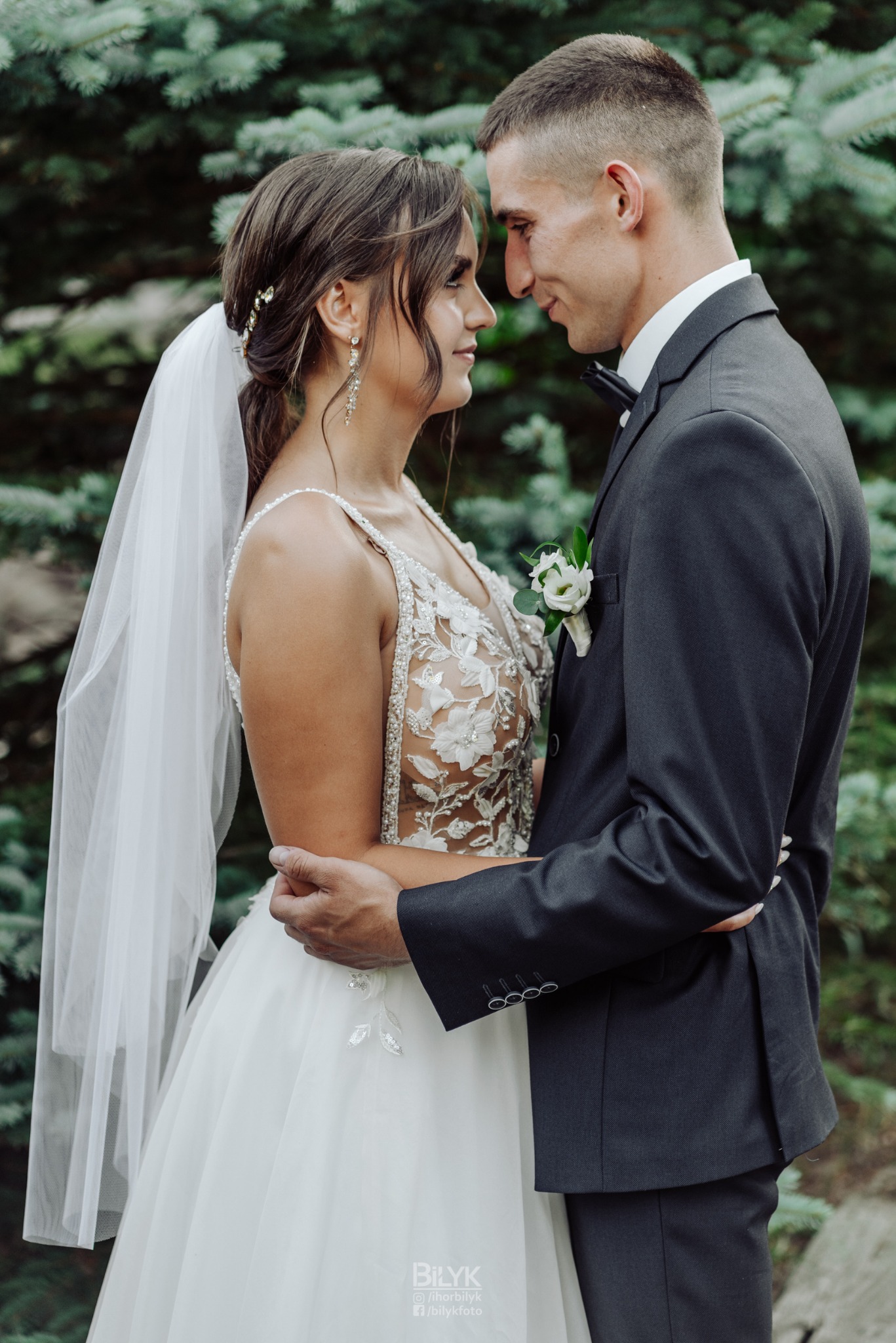 Para młoda w zbliżeniu: panna młoda w sukni z koronkowymi aplikacjami i welonem, pan młody w ciemnym garniturze z butonierką, czule obejmując się na tle zielonych świerków.